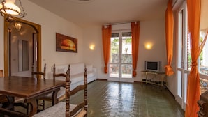 A dining area with a wooden table and chairs, a sofa, a painting, a television, and a door with curtains. - Lovely little house a few meters from the beach of the romantic Manerba (Manerba)