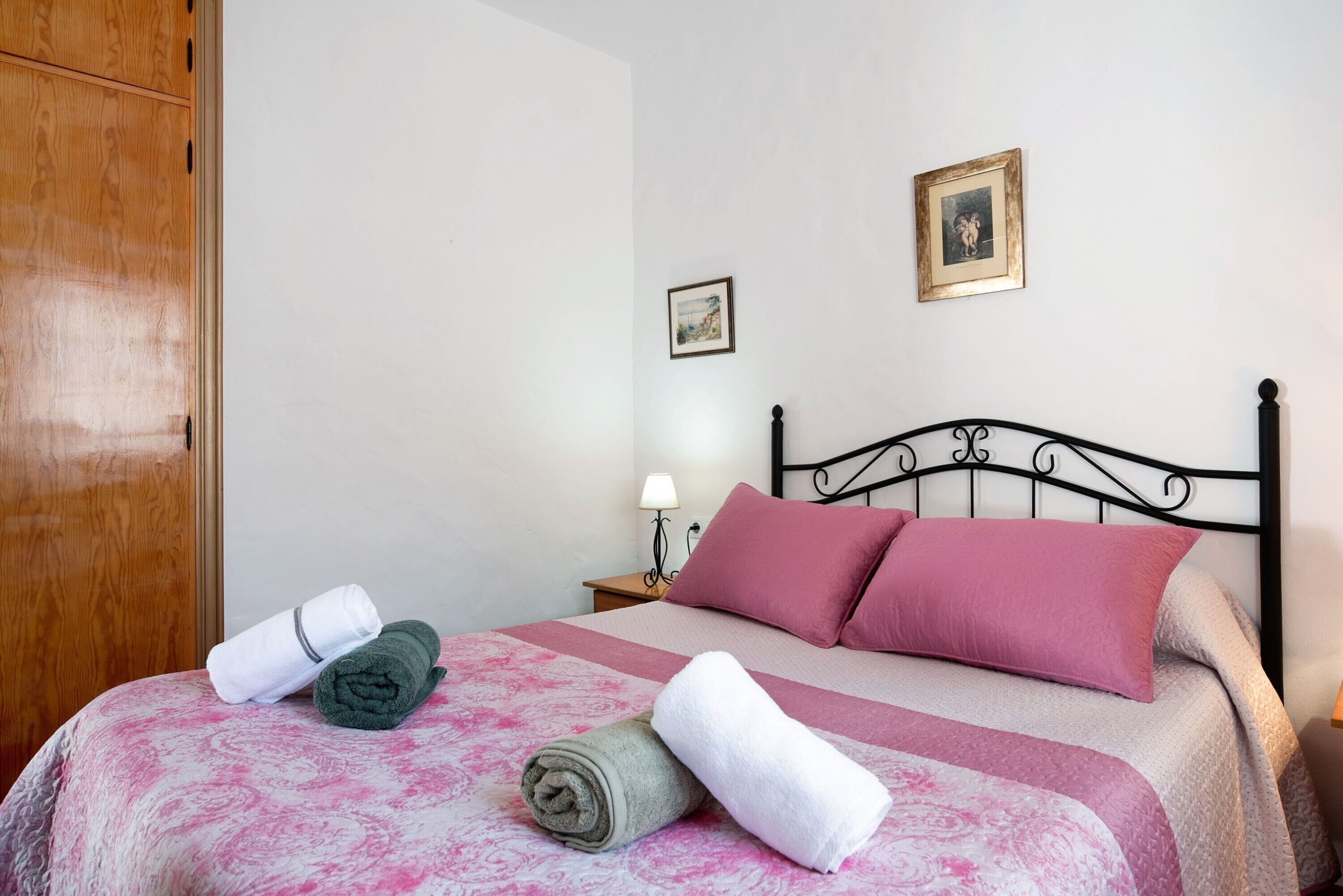 A neatly made bed with pink and white bedding, a wooden wardrobe, and a nightstand with a lamp.
