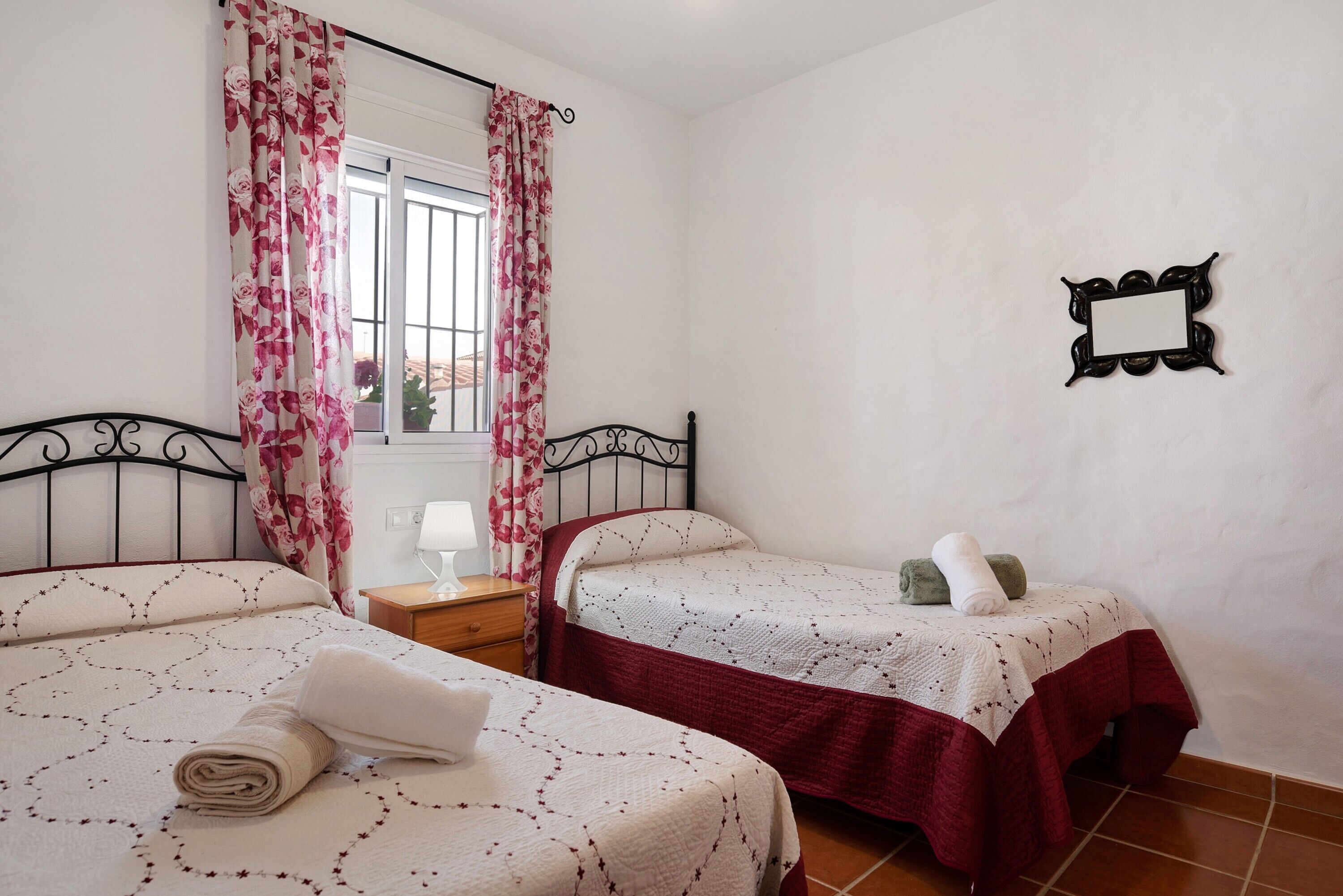 A bedroom with two beds, a nightstand, a lamp, a window with curtains, and a wall-mounted mirror.