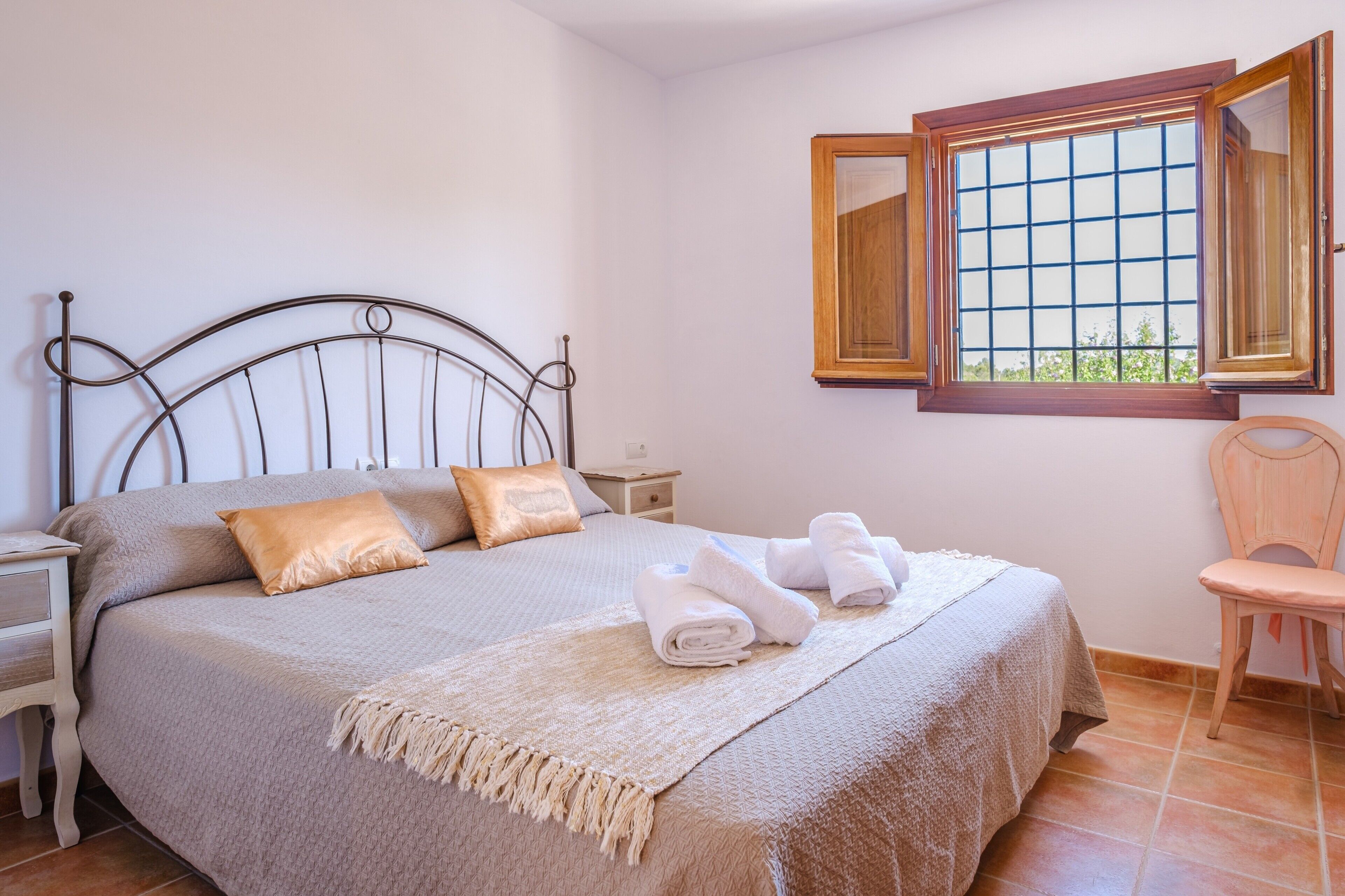 Een opgemaakt bed met hoofdbord, een houten stoel, een nachtkastje en een raam met houten kozijnen.