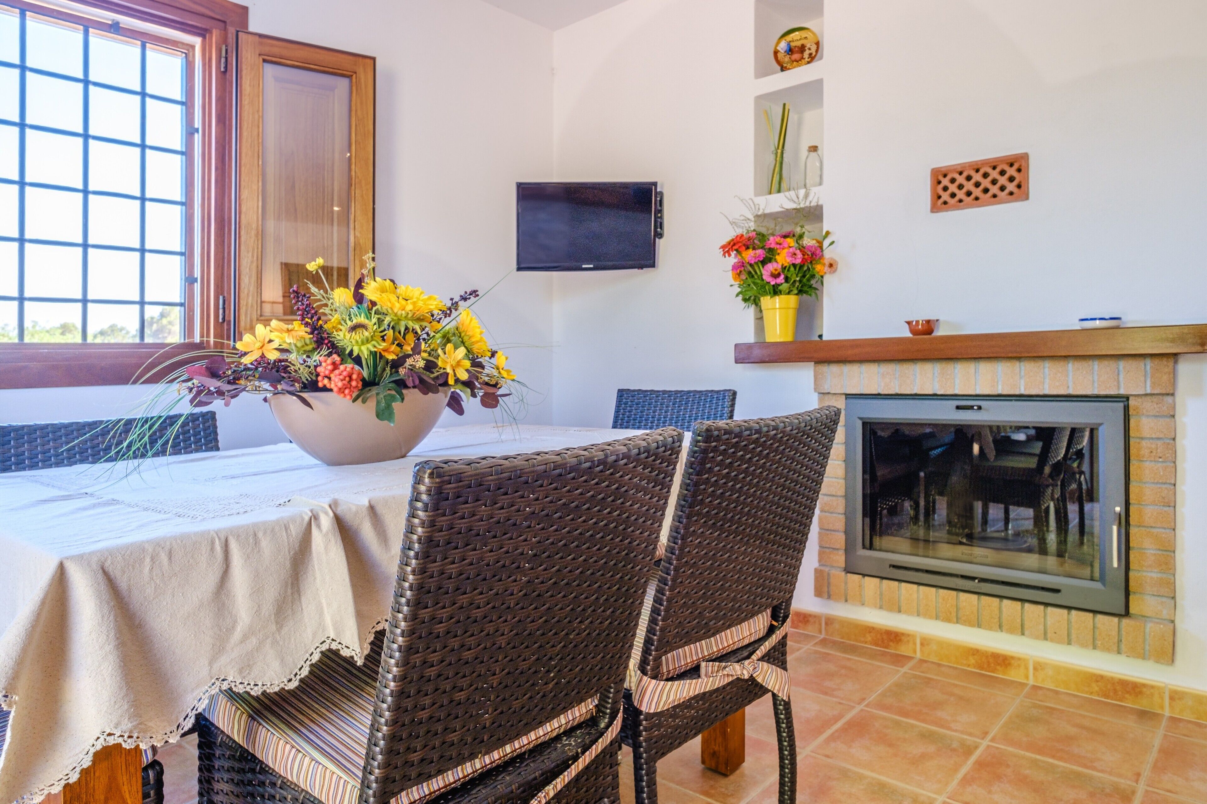 Un área de comedor con sillas de mimbre, una mesa con mantel, un florero, un televisor montado en la pared y una chimenea.
