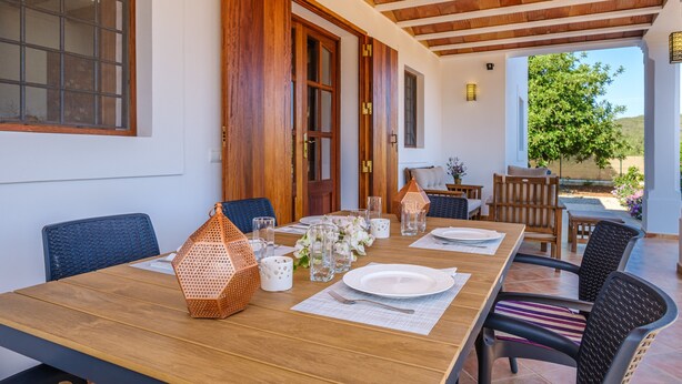 Een eethoek met een houten tafel voor vier personen, omringd door stoelen, met uitzicht op een tuin en een gebouw op de achtergrond.