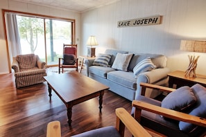 A living room with a wooden coffee table, a blue sofa with striped pillows, a red armchair, and a wooden side table with a lamp. A sign on the wall reads "LAKE JOSEPH".