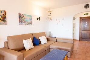 Een beige sofa met kussens, een klein tafeltje, wanddecoratie, een wandklok, een lamp, een hangende windgong, en een deur met de tekst 'SALIDA'.