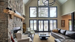 A spacious living room with a stone fireplace, large windows, a leather sofa, and a fireplace mantel. - Beautiful open-concept cottage on Moon River! 10 Minute walk to Bala! (Bala)