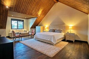 Un dormitorio con techo inclinado, cama con ropa de cama blanca, mesita de noche con lámpara, cómoda y ventana con vista a árboles.