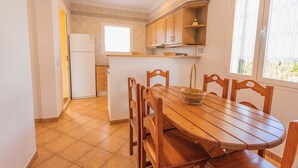 A kitchen with wooden cabinets, a white refrigerator, a dining table with chairs, and a window with a view. - Vacation Home "Villa Llebeig" Near the Beach with Sea View and Pool (San Jaime Mediterráneo)