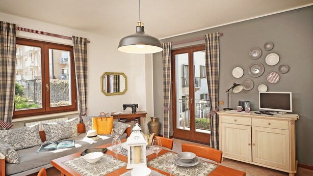 A dining area with a wooden table set for four, a sofa with cushions, a television on a cabinet, and a view through the window.