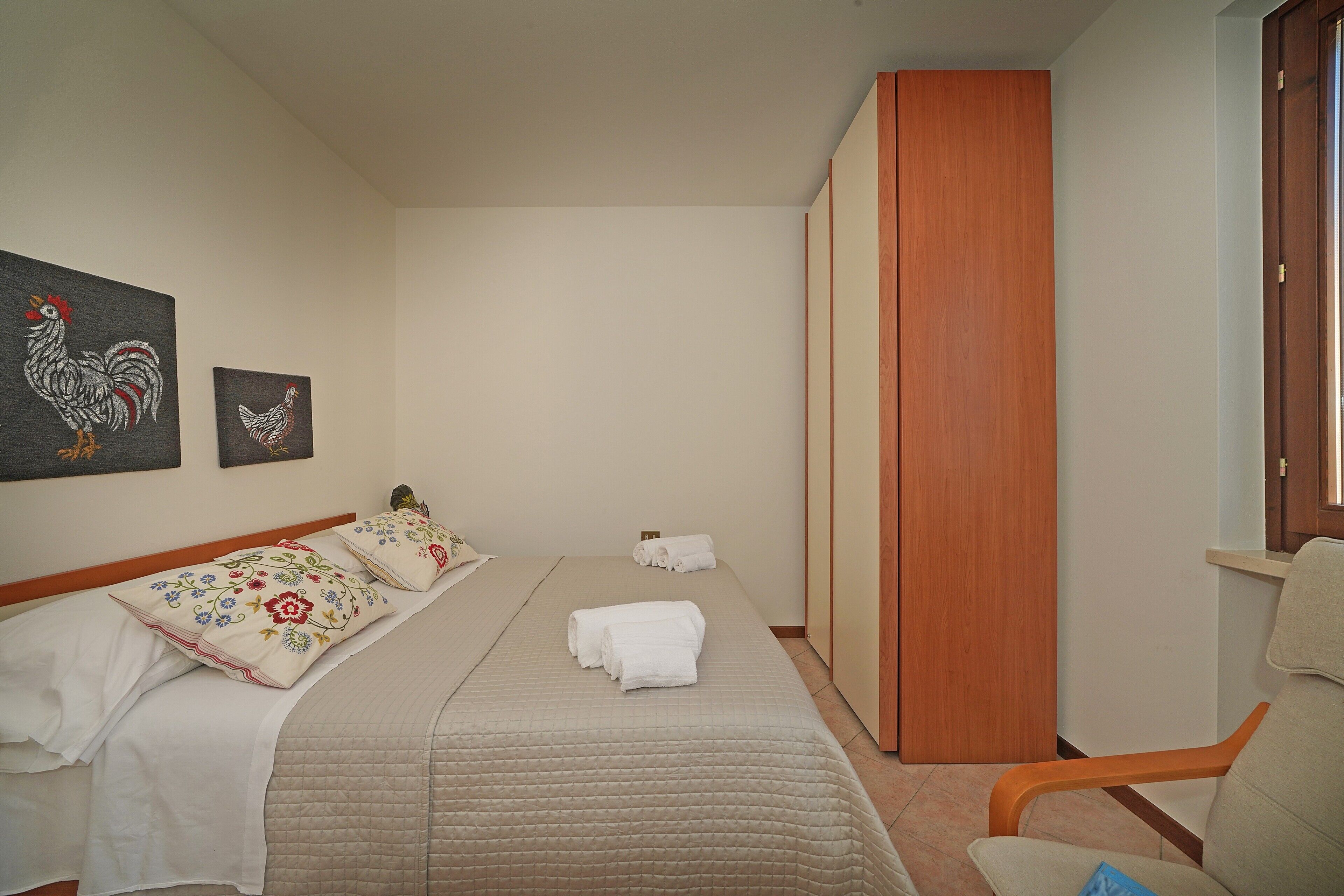 A hotel room with a bed, a chair, a wooden wardrobe, and two wall-mounted artworks.