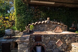 A stone-built outdoor kitchen with a built-in oven, surrounded by greenery and a thatched roof.
