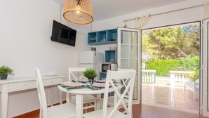 A compact kitchen with white cabinetry, a built-in TV, and a dining area with white chairs and a table set for two. - Beautiful apartment with porch, pool, garden and Wi-Fi (Son Xoriguer)