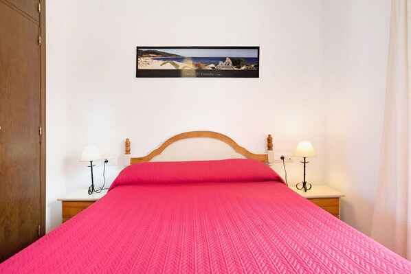 A bed with a red bedspread, two bedside lamps, a wooden headboard, and a framed picture of a beach scene on the wall.