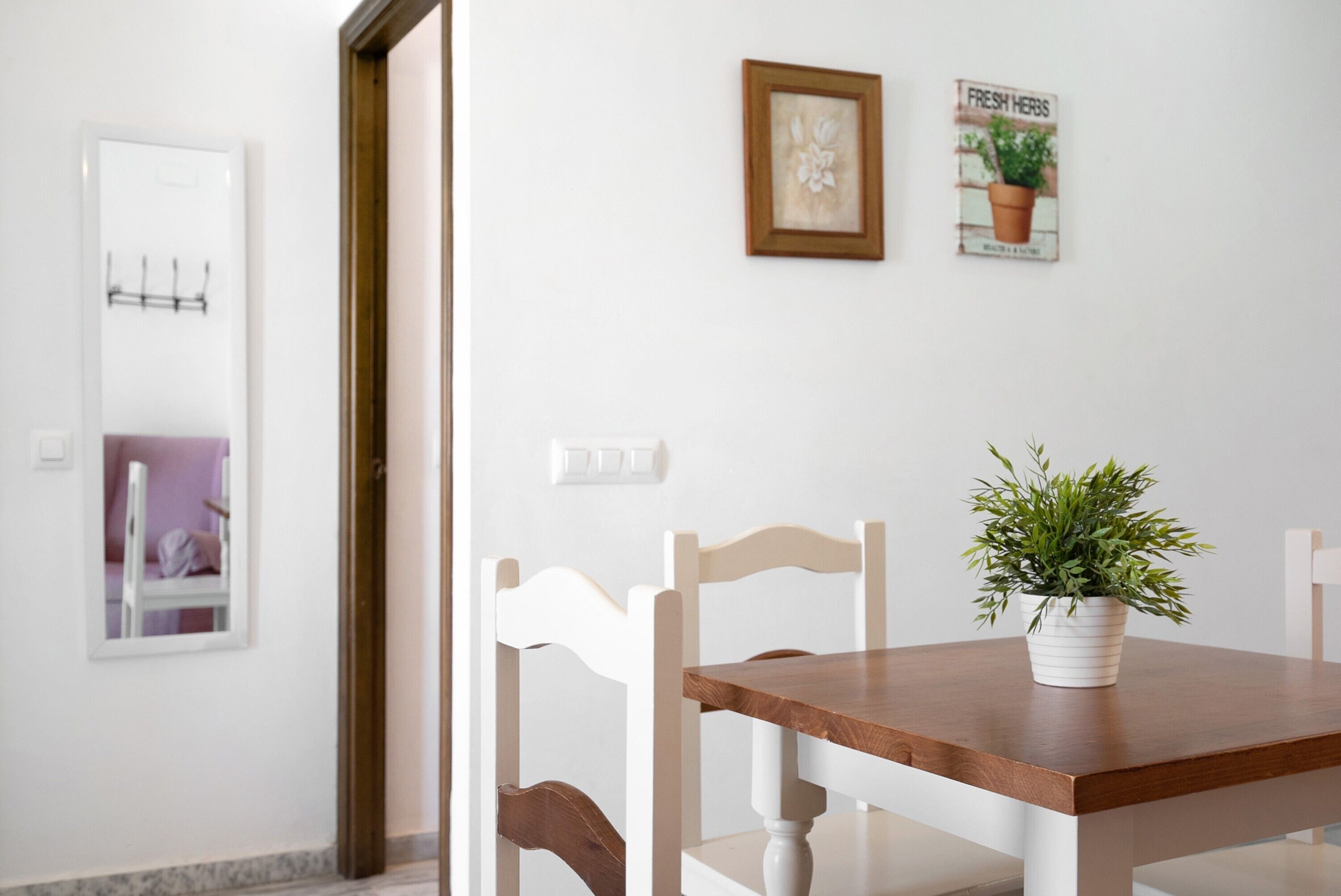 Un coin repas avec une table en bois et des chaises blanches, une plante en pot posée sur la table et un interrupteur fixé au mur.