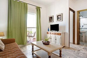 Una sala de estar con un sofá, una mesa con una cesta, un televisor sobre un mueble, y un baño visible a través de una puerta abierta.