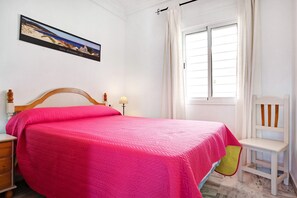 Un dormitorio con cama, mesita de noche, silla, ventana con cortinas e imagen colgada en la pared.