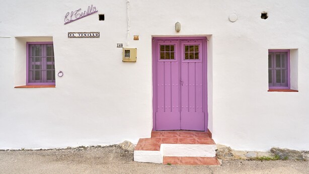 A white building with a purple door, two windows with purple frames, and a small sign above the door. - Holiday Home 'Casa Rural El Tesillo' with Mountain View and Wi-Fi (Canillas de Aceituno)