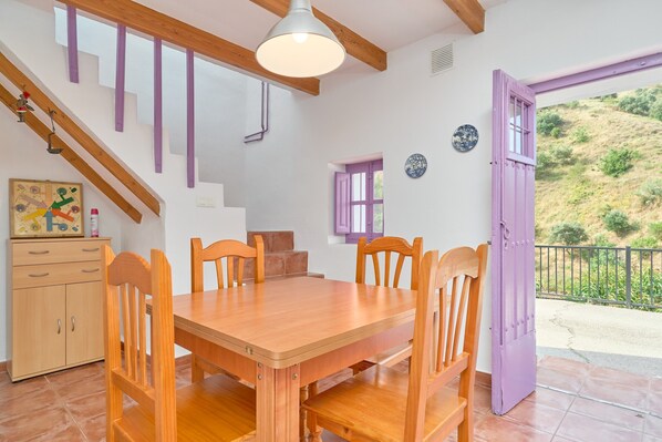 A dining area with a wooden table and chairs, a staircase with purple railings, and a view of a hillside through a door.