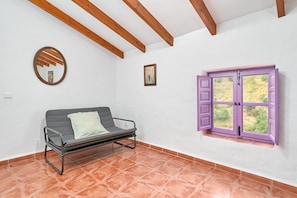 A room with a metal-framed sofa, a window with purple shutters, a wooden ceiling beam, and a tiled floor.