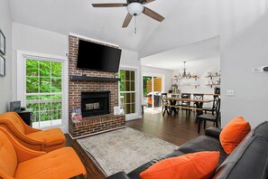 Ein Wohnzimmer mit Kamin, orangefarbenen Sesseln, einem grauen Sofa und einem Essbereich mit Tisch und Stühlen.