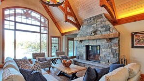 A cozy living room with a stone fireplace, wooden beams, and large windows. - Gorgeous 7 Bed, 6 Bath Family Cottage on Lake Rosseau! Muskoka Room! Pool Table! (Port Carling)