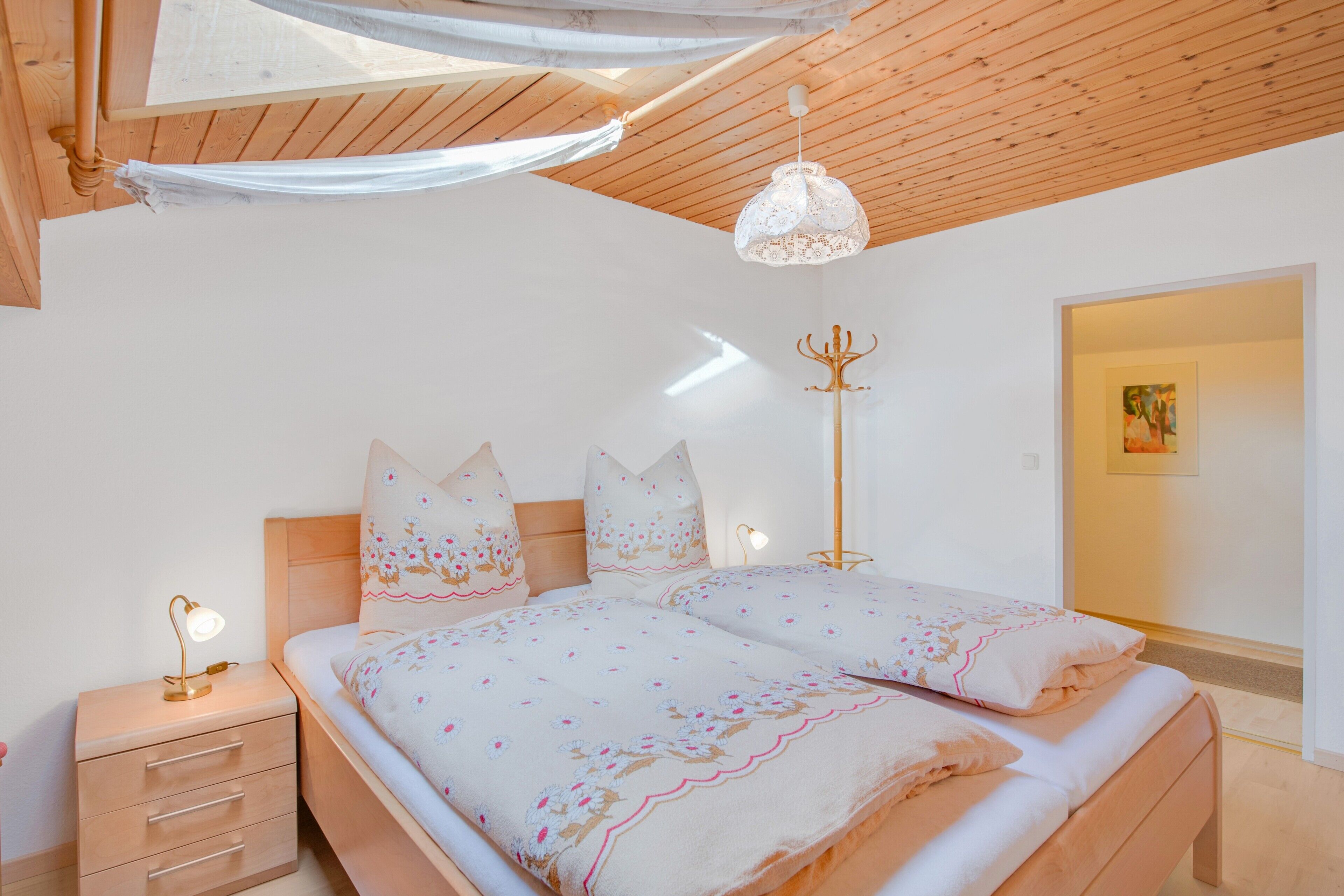 Ein Schlafzimmer mit Holzbalkendecke, einem Bett mit floralem Bettzeug, einem Nachttisch mit Lampe und einem Kleiderständer.