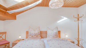 Quarto com duas camas, cabeceira de madeira, mesa de cabeceira com abajur, porta-roupas e janela com cortina branca.