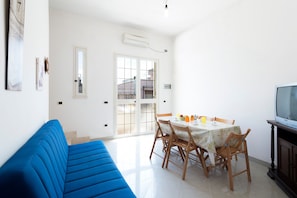 A bright living room with a blue sofa, a dining table with chairs, a TV, and a large window.