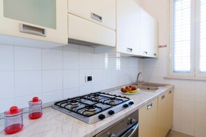 Una cocina con armarios blancos, estufa a gas, lavamanos y una ventana con persianas.