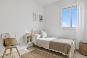 A bedroom with a bed, a chair, a bedside table with books, a window, and a wall with two framed pictures.