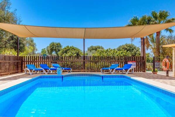 A swimming pool with blue tiles, surrounded by blue lounge chairs, a wooden fence, and palm trees. - Charming Finca with Pool, Garden, Terrace, Air Conditioning & Wi-Fi (Santanyí)