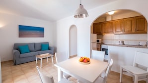 A modern kitchen with wooden cabinets, a white dining table with chairs, and a fruit bowl. - Modern apartment in complex with pool - Girasols Gardens Sup (Son Bou)