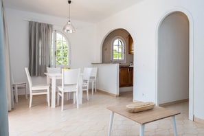 A dining area with a table and chairs, a kitchen with an archway, and a window with curtains.