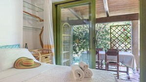 A bedroom with a bed, a hat, and towels on it, a chair, a table, and a view of greenery outside. - Cottage with Garden, Terrace, Air Conditioning, and Shared Pool (Porto Rotondo)