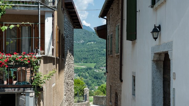 Una calle estrecha con edificios de piedra, un balcón adornado con flores rojas y una vista de la montaña en la distancia.