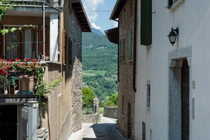 A narrow street with stone buildings, a balcony with red flowers, and a mountain view in the distance. - Bright and comfortable - Appartamento Acqua (Tignale)