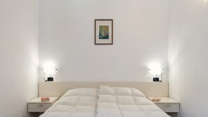 Un dormitorio con cama, dos mesitas de noche, una mesita auxiliar, un cuadro enmarcado en la pared y dos lámparas de pared.