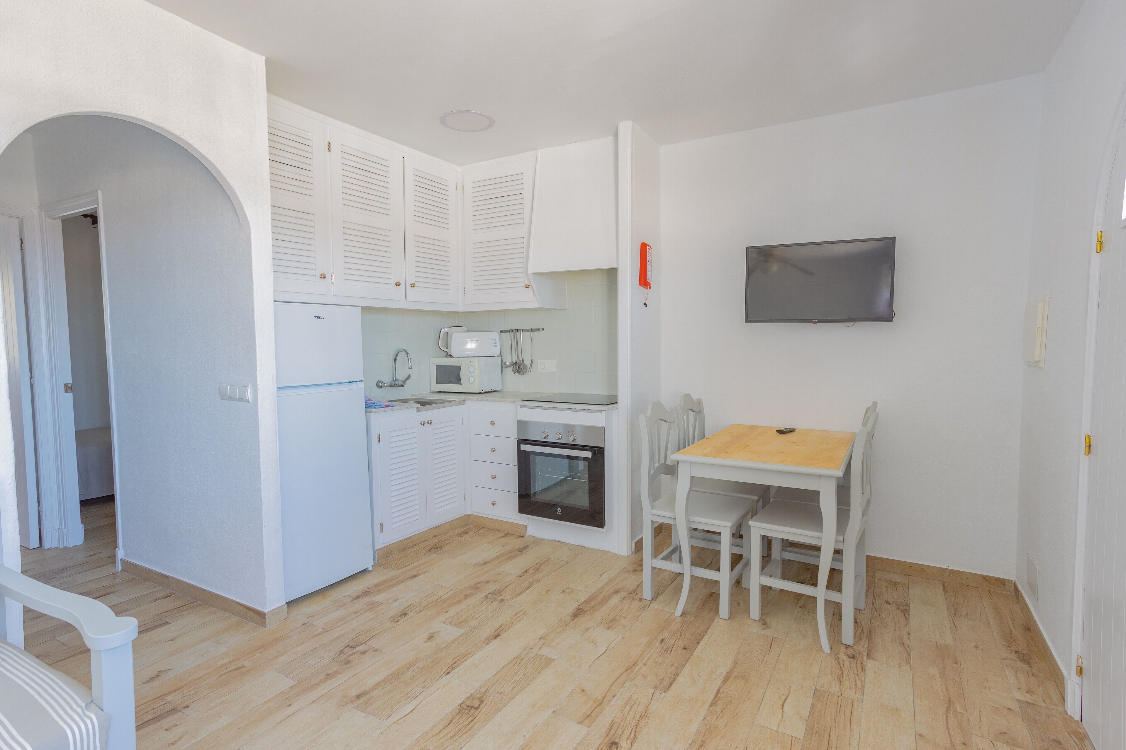 Een kleine keuken met witte kastjes, een magnetron, een waterkoker en een kleine oven. Er is een houten eettafel met stoelen en een flatscreen televisie aan de muur bevestigd.