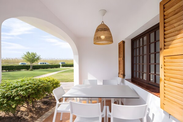 A patio with a table and chairs, a hanging light fixture, a window with wooden shutters, and a view of a landscaped garden.