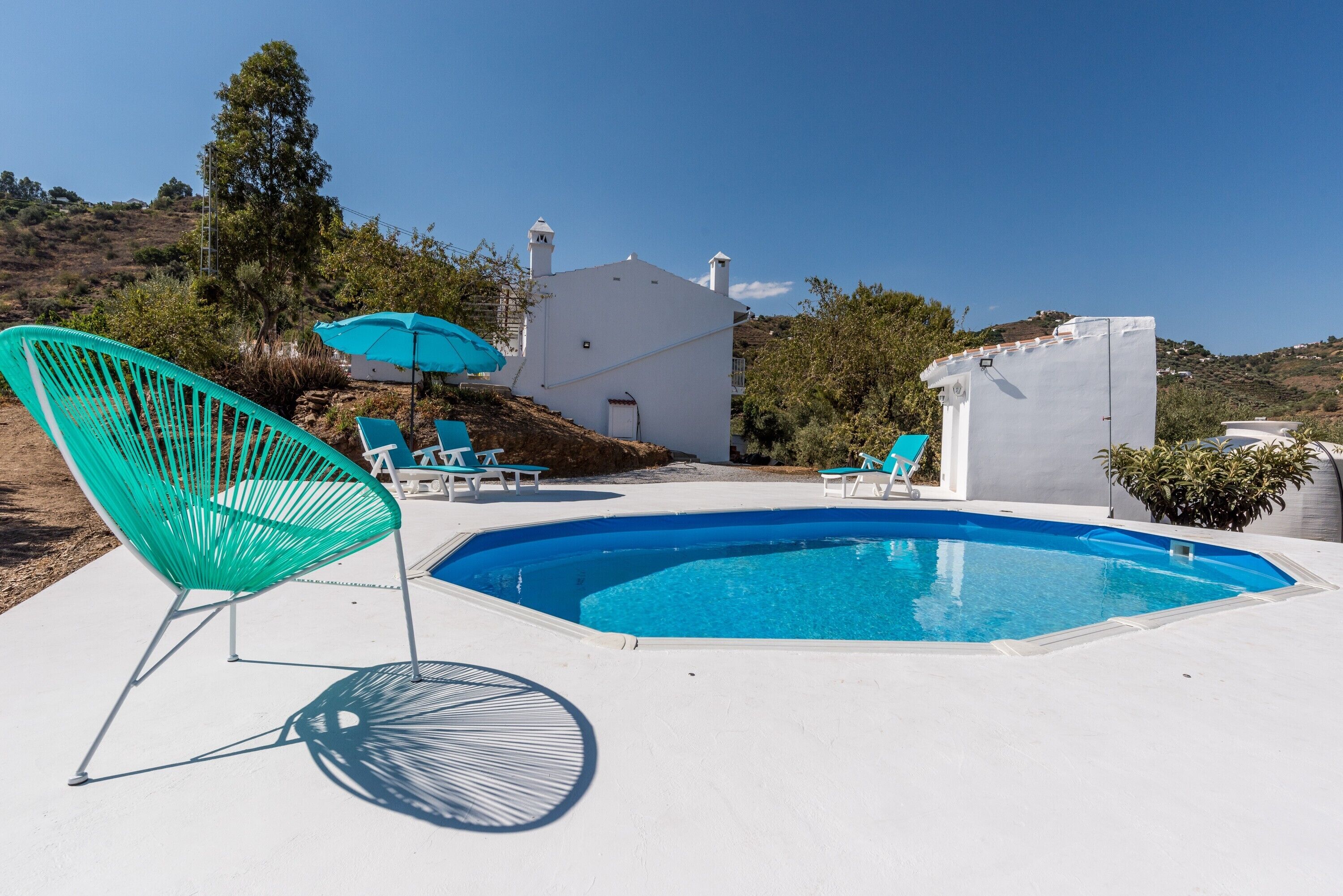 Una piscina con sillones de playa y sombrillas, rodeada por un edificio blanco con techo inclinado y chimeneas, ubicada en una ladera.