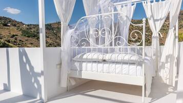Ein weißes Metallbett mit Baldachin, weiße Wände und Blick auf eine hügelige Landschaft.
