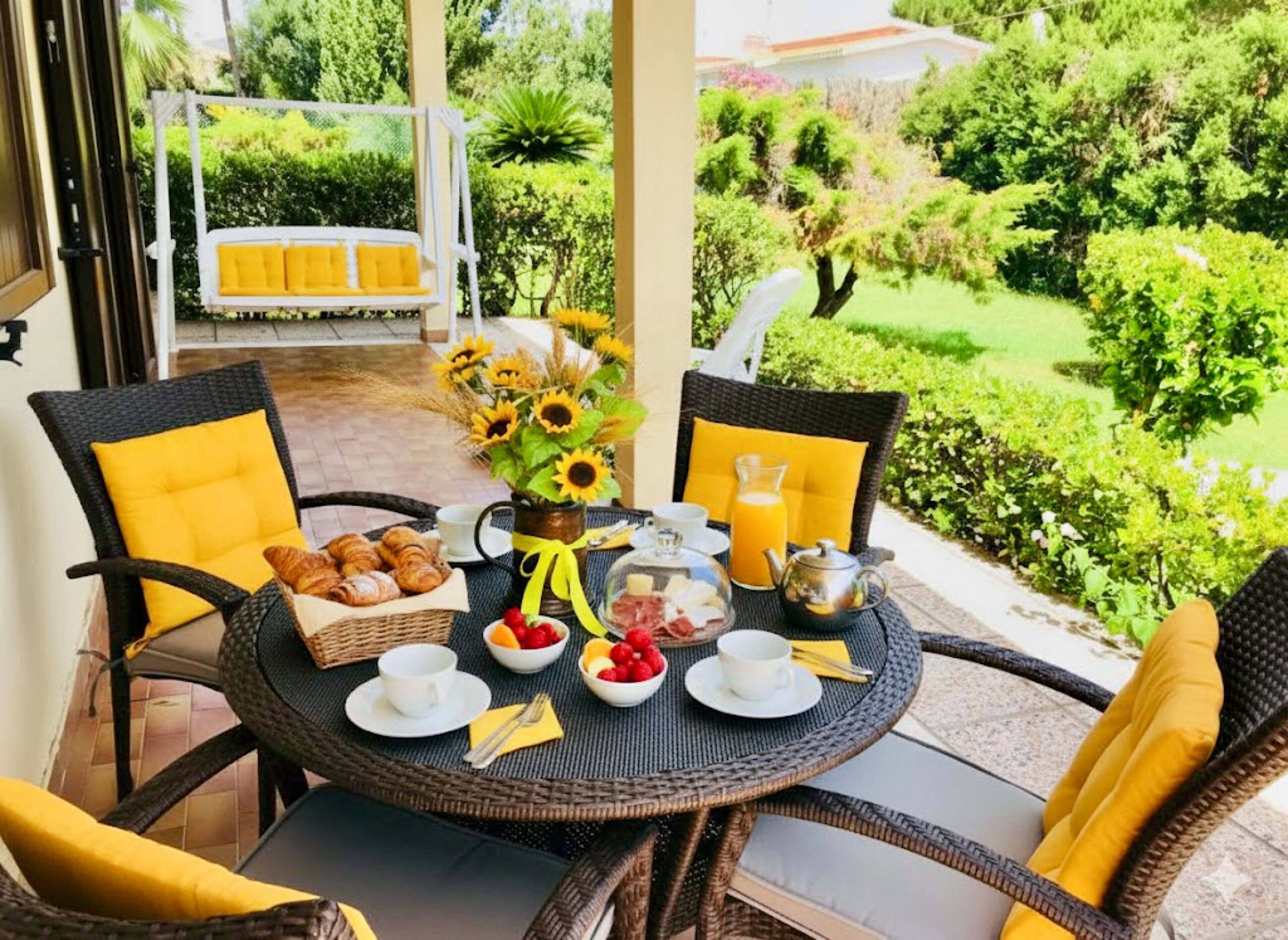Ein runder Tisch mit Korbstühlen, einem Korb voller Croissants, einer Vase mit Sonnenblumen und Frühstücksleckereien.