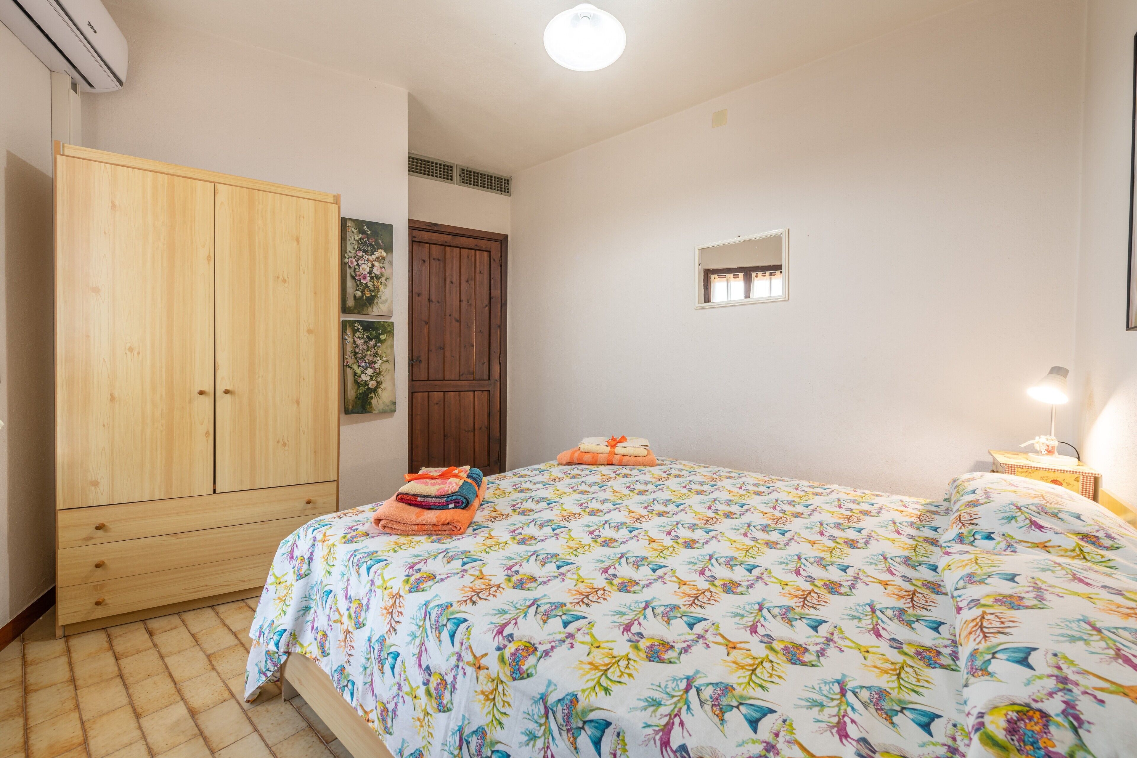 Ein Schlafzimmer mit Holzschrank, Bett mit Blumenmuster und Nachttisch mit Lampe, sowie ein kleines Fenster.