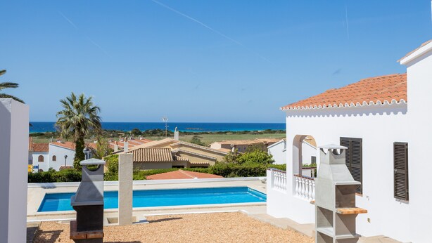 A pool area with a barbecue grill, a view of the sea, and a white building with a tiled roof. - Family Villa with Sea Views (San Jaime Mediterráneo)