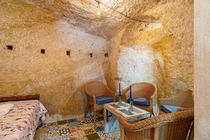 A room with a bed, two wicker chairs, a small table with a candle, and a tiled floor.