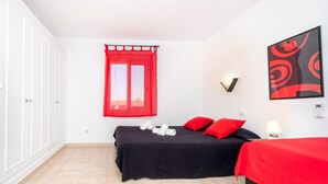 A bedroom with a bed, a window with red curtains, a wall-mounted light, and a painting.