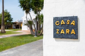 Ein Schild mit dem Schriftzug 'CASA ZARA' an einer weißen Wand, ein Weg und etwas Grün.