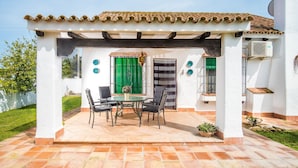 Une terrasse couverte avec une table et des chaises, une porte verte et un mur blanc surmonté d’un toit en tuiles.