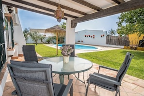 Eine überdachte Terrasse mit einem Glastisch und Stühlen, einem hängenden Laternenlicht und Blick auf einen Swimmingpool sowie ein Haus mit weißer Fassade und roten Blumen.