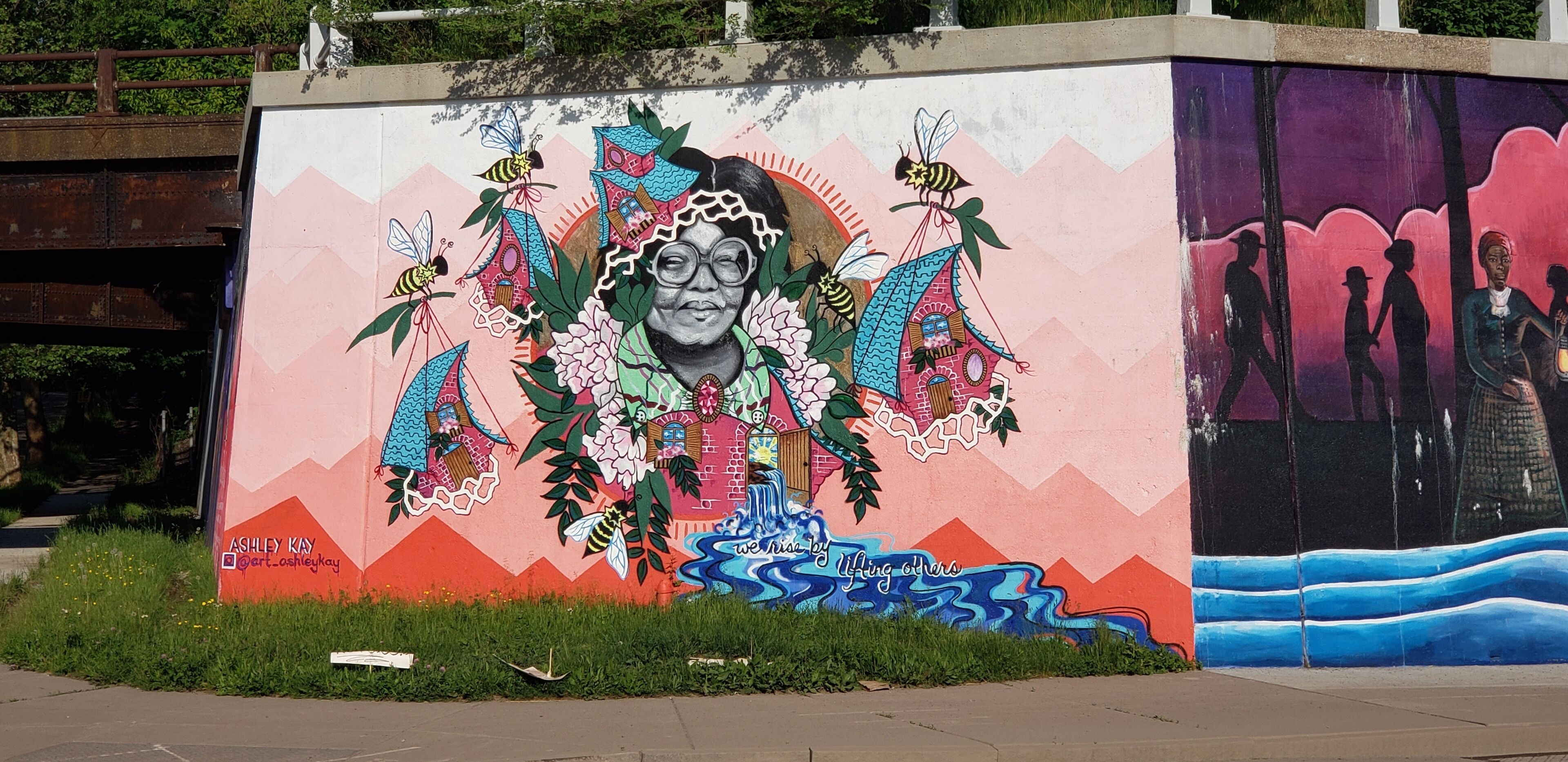 A mural with a central figure surrounded by bees and flowers, and the phrase "we rise by lifting others" at the bottom.