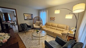 Un salon comprenant un canapé, deux fauteuils, une table basse et, au fond, un coin repas.